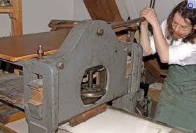 Der Steindrucker steht an einer Lithographiepresse und drückt einen Hebel herunter. 