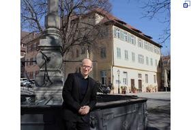 Der neue Museumsleiter steht am Brunnen auf dem Hafenmarkt. Im Hintergrund befindet sich das Stadtmuseum im Gelben Haus. Hansjörg Albrecht ist etwa 50 Jahre alt, hat kurze Haare und eine Brille und lächelt in die Kamera. Er trägt ein dunkles Sakko und ein dunkles Hemd.