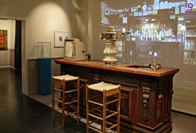 Ausstellungsraum: Ein Kneipentresen mit dekorativer Zapfanlage, davor zwei Barhocker. Dahinter ist eine Kneipenwand mit Flaschen und so weiter projiziert.