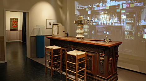 Ausstellungsraum: Ein Kneipentresen mit dekorativer Zapfanlage, davor zwei Barhocker. Dahinter ist eine Kneipenwand mit Flaschen und so weiter projiziert.