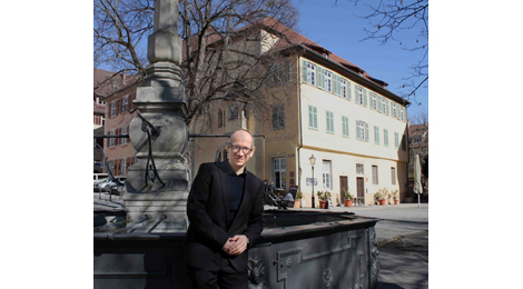 Der neue Museumsleiter steht am Brunnen auf dem Hafenmarkt. Im Hintergrund befindet sich das Stadtmuseum im Gelben Haus. Hansjörg Albrecht ist etwa 50 Jahre alt, hat kurze Haare und eine Brille und lächelt in die Kamera. Er trägt ein dunkles Sakko und ein dunkles Hemd.