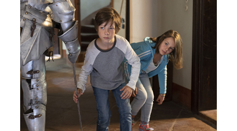 Foto aus dem Film Max und die wilde Sieben: Ein Junge und ein Mädchen schleichen vorsichtig um eine Ritterrüstung herum, die als Dekoration in einem großen Raum steht. 