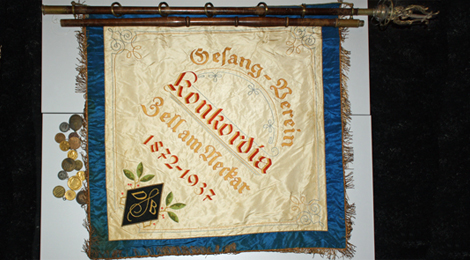 Auf einem Tisch liegt ausgebreitet die Vereinsfahne. Sie ist aufwändig geschmückt und bestickt mit den Worten "Gesang-Verein Konkordia Zell am Neckar 1872-1937" .